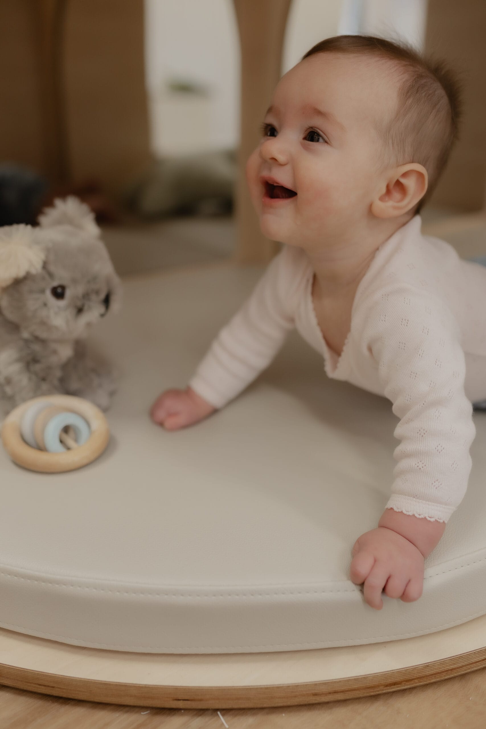 A baby crawling and laughing on a soft round mat with a plush toy and wooden rattle