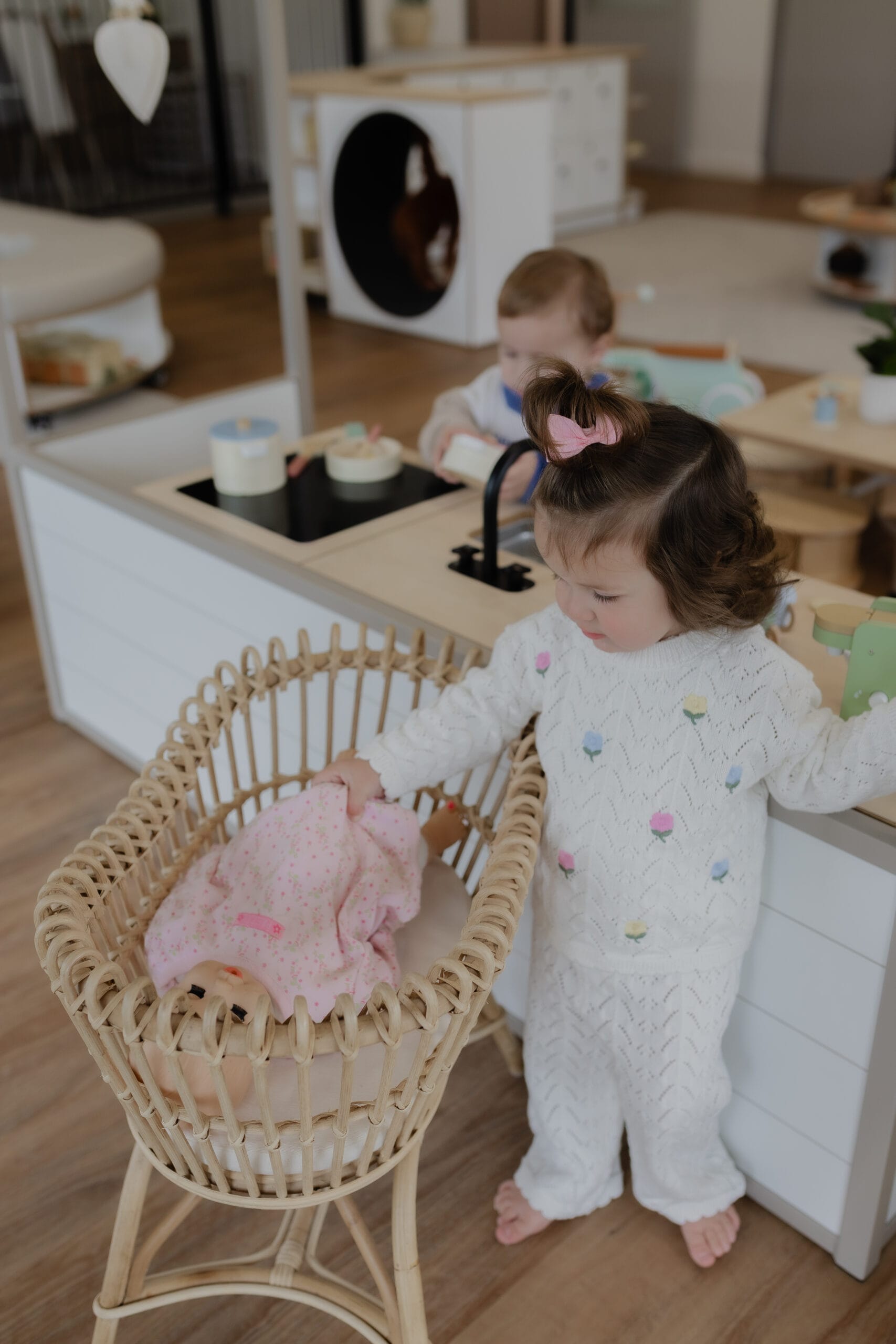 A girl playing with a doll in a cradle basket in the play kitchen area