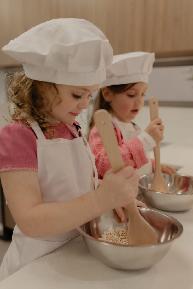 Two girls wearing chef hats mixing ingredients in bowls with wooden spoons