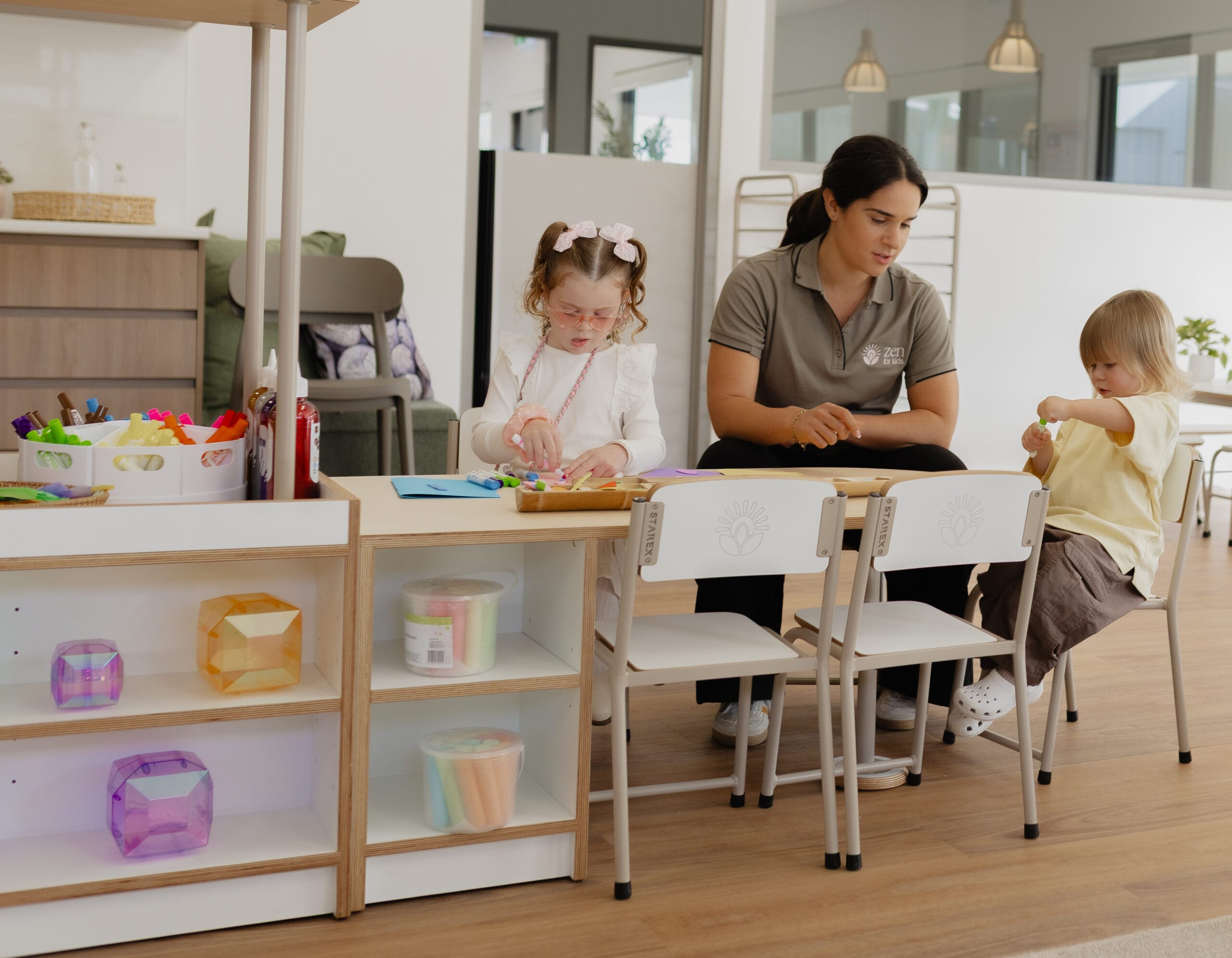 An educator sitting between two children doing a craft activity at a table in the Moon Room