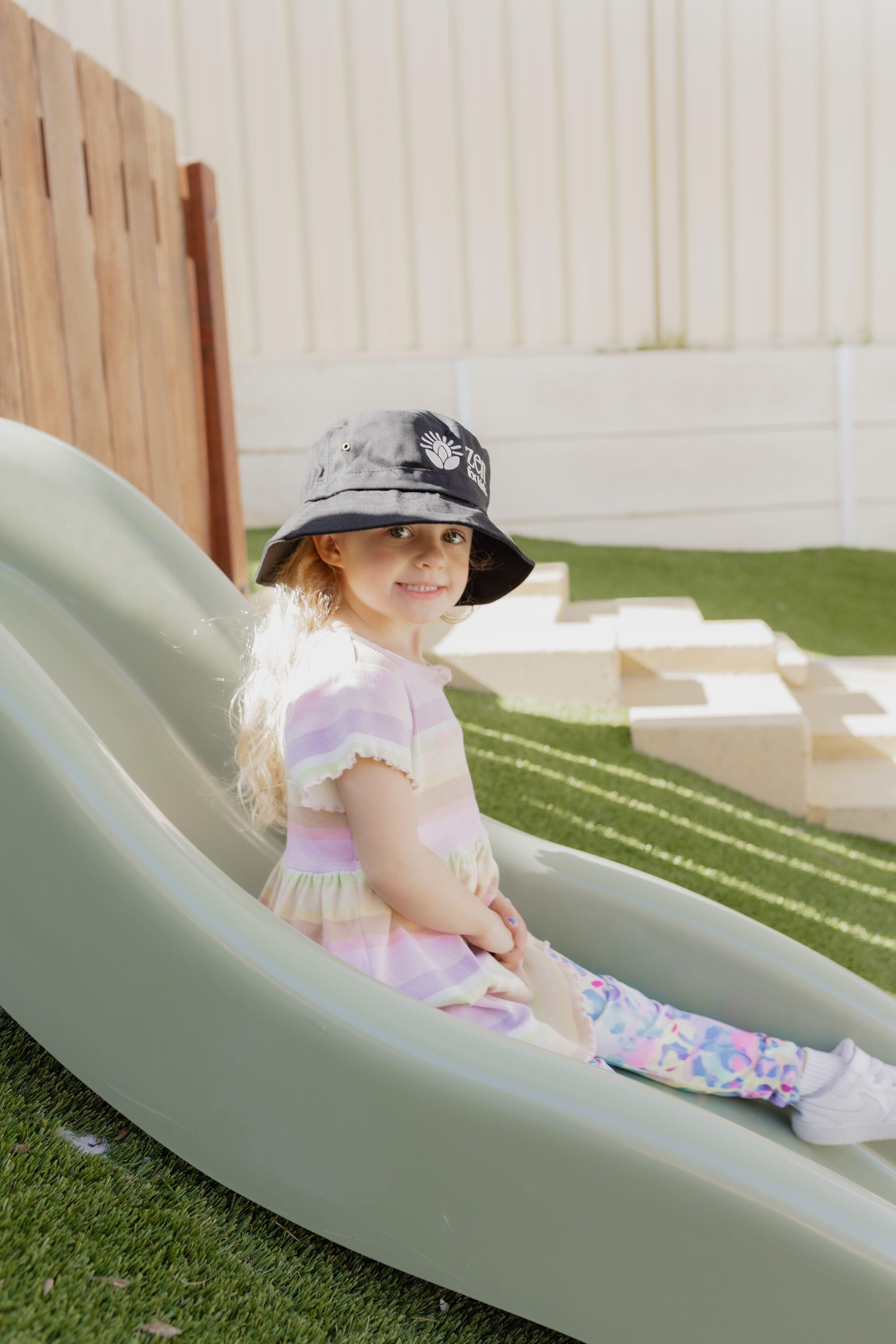 A girl wearing a bucket hat sitting on an outdoor slide in the play yard
