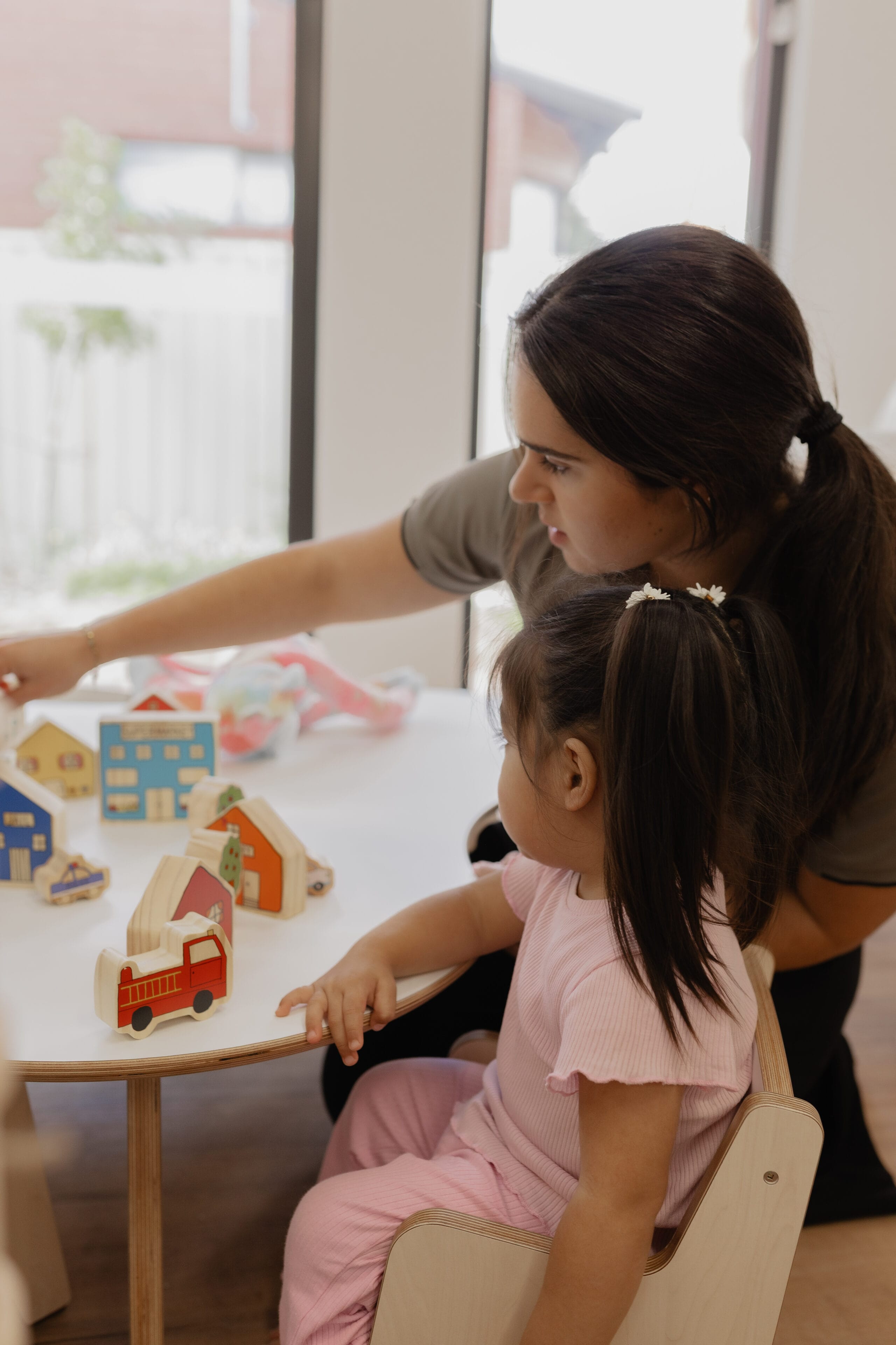 An educator showing a young girl wooden toy houses and vehicles at a table