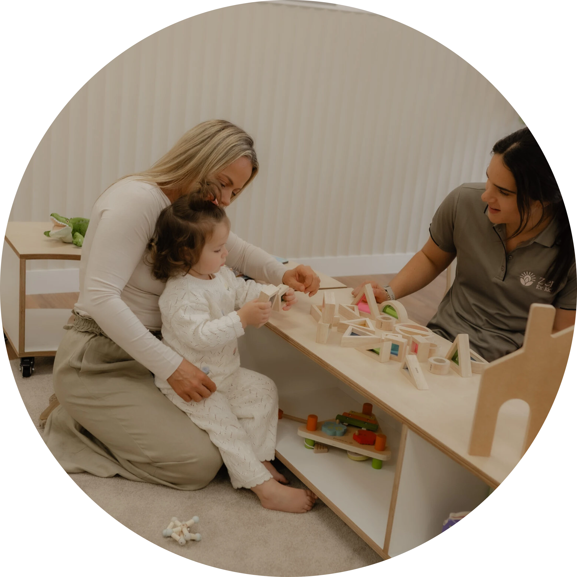 Two educators sitting on the floor with a toddler playing at a low shelf