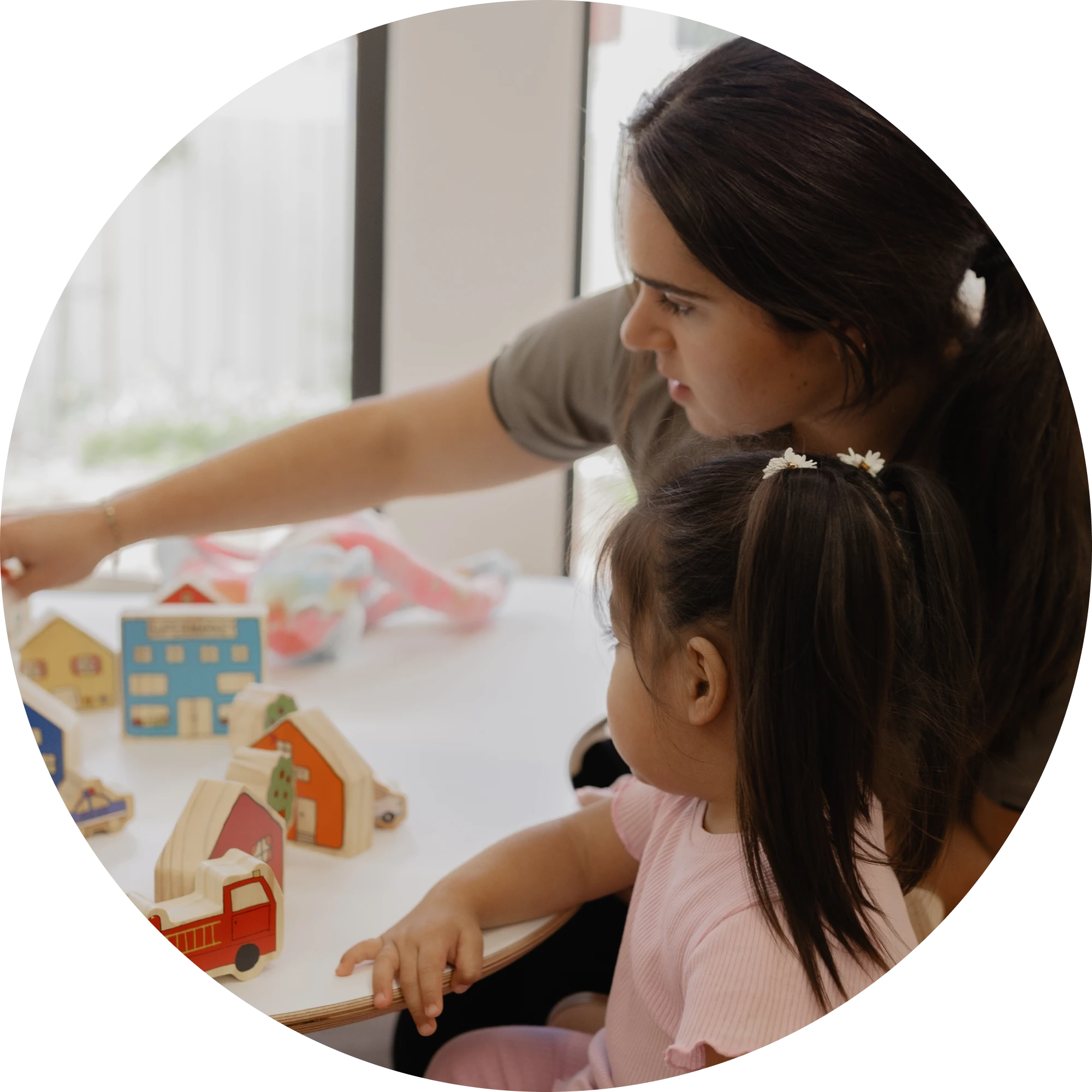 An educator pointing to a wooden toy village while sitting with a child