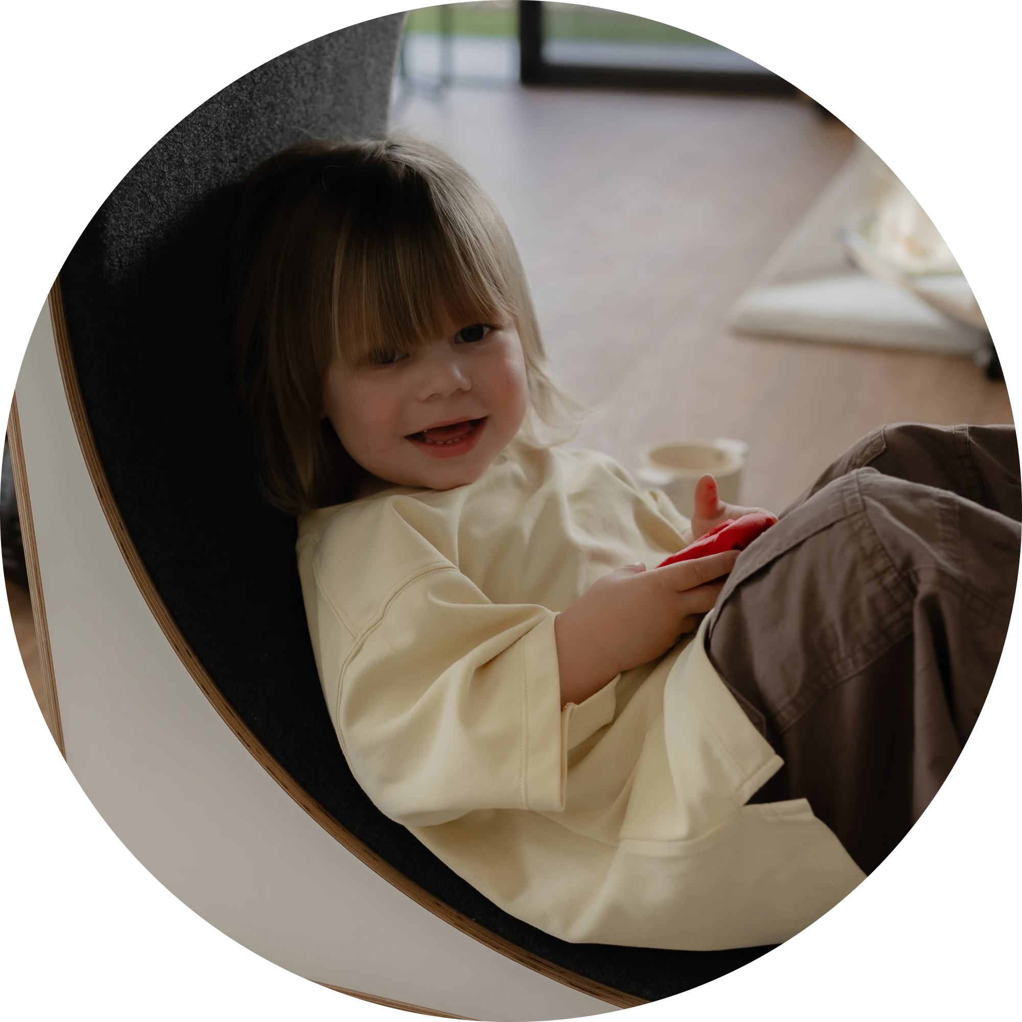 A smiling toddler sitting with an educator in a round chair, holding a toy phone